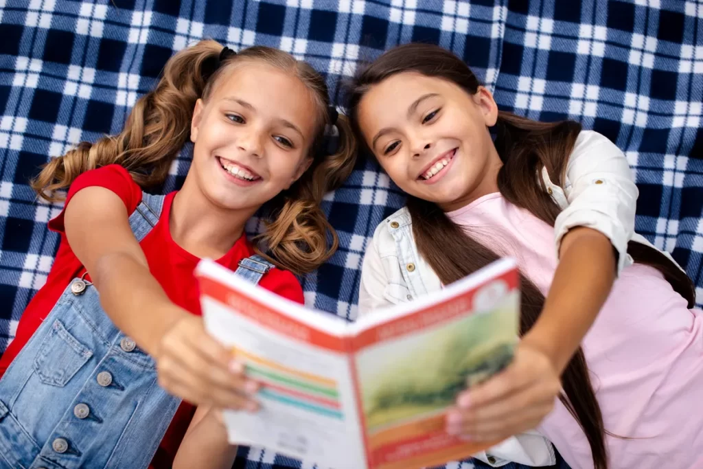 Duas meninas pré-adolescentes, deitadas em uma manta xadrez azul marinho e branca, estão sorrindo para a câmera enquanto seguram e leem juntas um livro de capa vermelha. A menina da esquerda está de macacão jeans e blusa vermelha e a da direita está de blusa rosa e jaqueta branca, em um momento de amizade e aprendizado compartilhado.