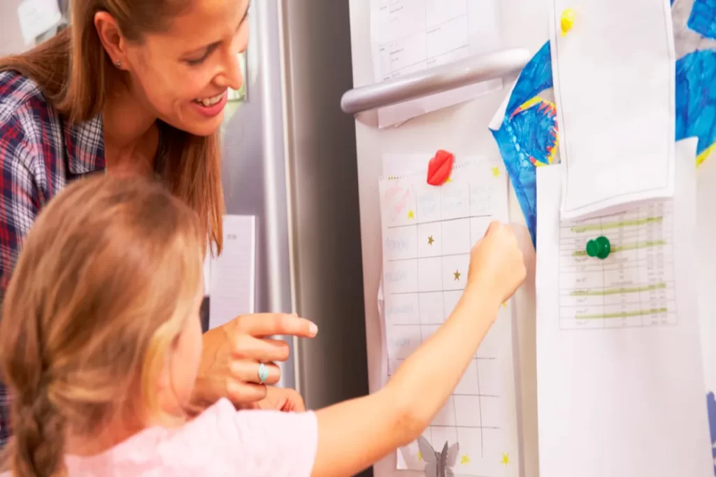 Uma mãe sorridente, vestindo uma camisa xadrez, e sua filha pequena, com cabelo loiro, estão em frente a uma geladeira branca. A menina está colocando uma estrela dourada adesiva em um quadro de incentivo semanal ou mensal preso à geladeira, que lista tarefas ou metas. A mãe aponta para a tabela, reforçando o uso do quadro de recompensas para o comportamento infantil.