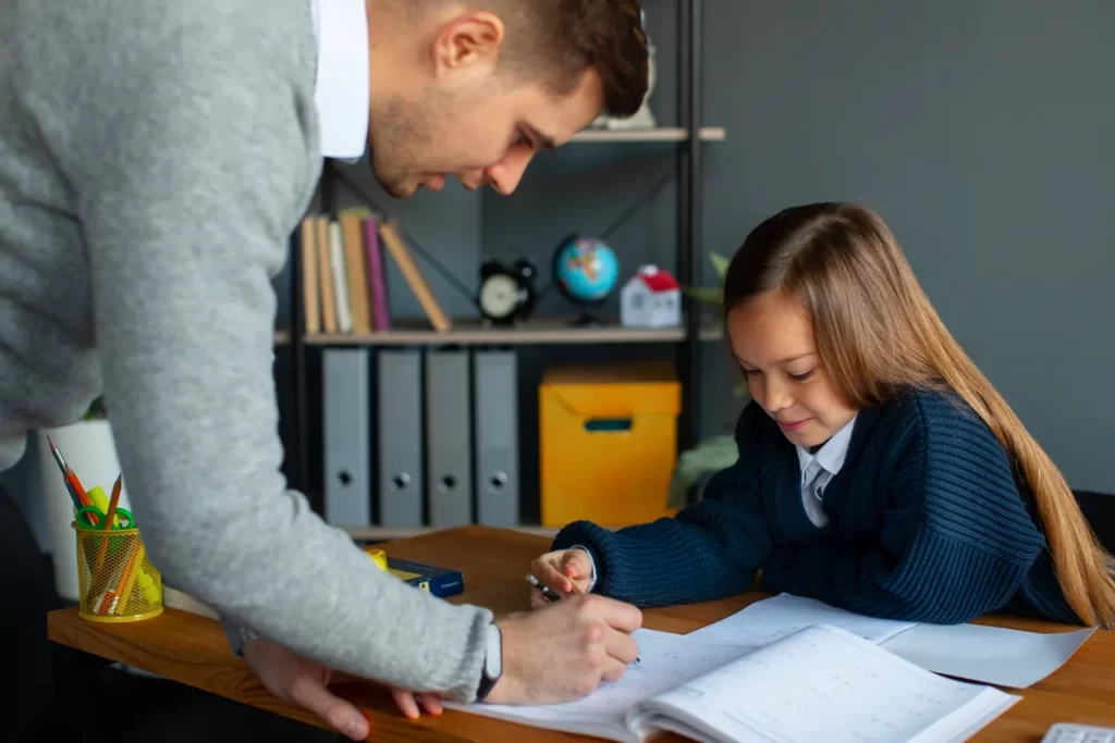 Um homem adulto de suéter cinza e camisa social branca inclina-se sobre uma mesa de madeira para auxiliar uma menina de cabelo castanho comprido e uniforme escolar azul marinho a escrever ou desenhar em um caderno aberto. O homem está segurando uma caneta, apontando para o caderno. A cena, em um escritório ou quarto, sugere a criação de regras, o estudo ou o dever de casa.