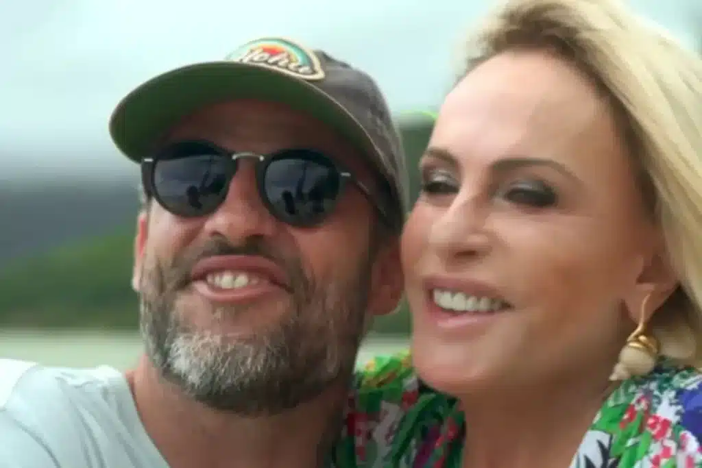 Duas pessoas adultas sorrindo lado a lado em um ambiente ao ar livre, uma delas usando boné e óculos escuros, e a outra com brincos e roupa colorida, transmitindo leveza e descontração