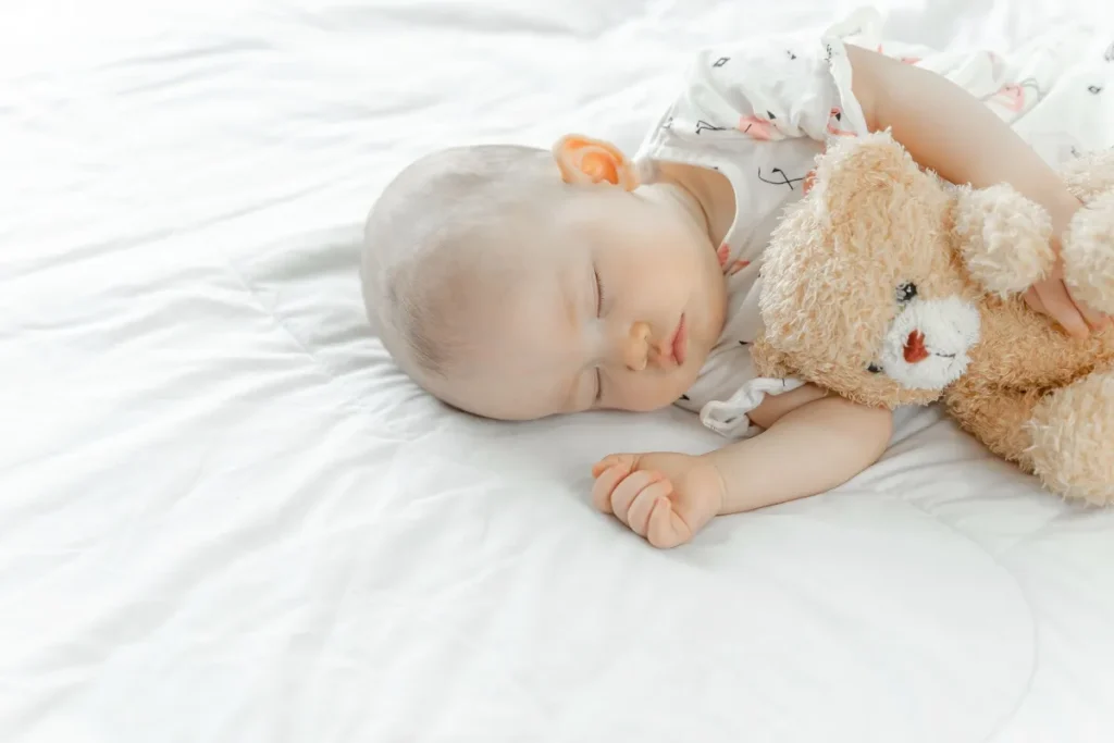 Bebê dormindo tranquilamente de lado em um colchão plano com lençol branco, abraçado a um ursinho de pelúcia marrom claro.