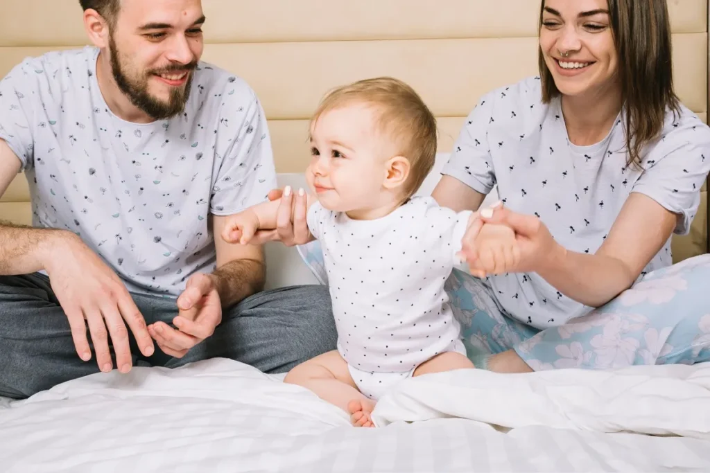 Um homem barbudo e uma mulher sorridente com piercing no nariz, ambos de pijama claro, sentados em uma cama branca e segurando as mãos de um bebê de body branco para ajudá-lo a ficar em pé.