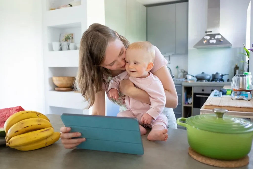 Mulher na cozinha com um bebê de macacão rosa no colo, olhando para a tela de um tablet com capa azul posicionado na bancada perto de bananas e uma panela verde.
