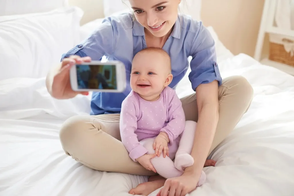 Mulher de camisa azul sentada em uma cama branca tirando uma selfie com o celular ao lado de um bebê sorridente de macacão rosa com bolinhas brancas.