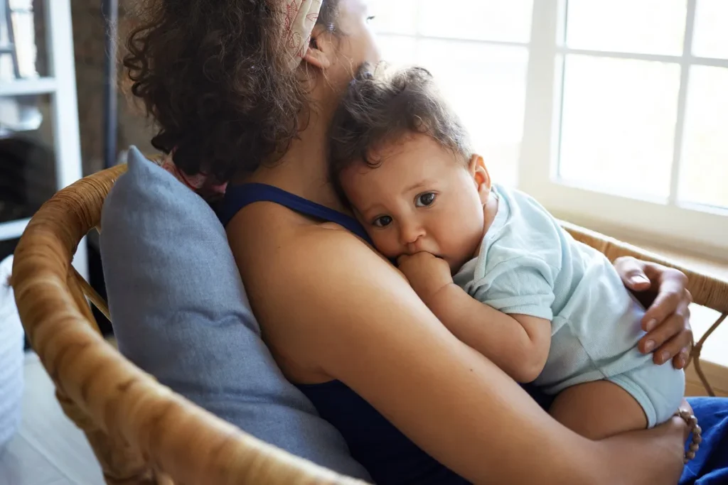 Mãe sorridente sentada em poltrona cinza, amamentando o bebê no colo enquanto apoia as costas em travesseiro de cabeceira.