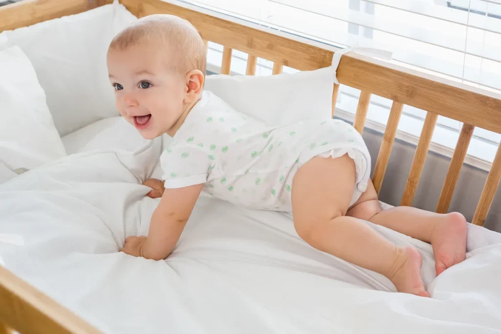 Bebê sorridente de pijama branco de bolinhas verdes engatinhando sobre um colchão firme dentro de um berço de madeira clara com protetores brancos acolchoados.