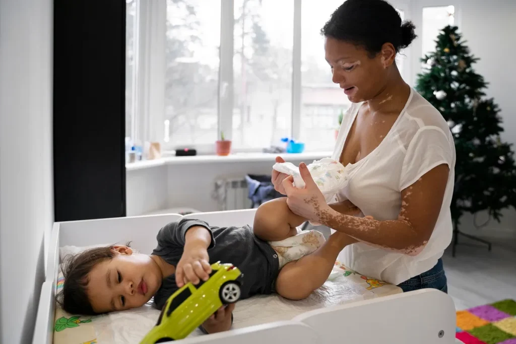 Mãe em pé trocando a fralda de um bebê mais crescido deitado no trocador, que está posicionado próximo a uma grande janela iluminada.
