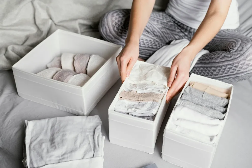 Mulher sentada organizando roupas dobradas em arquivo vertical dentro de caixas organizadoras brancas de tecido sobre uma cama cinza.