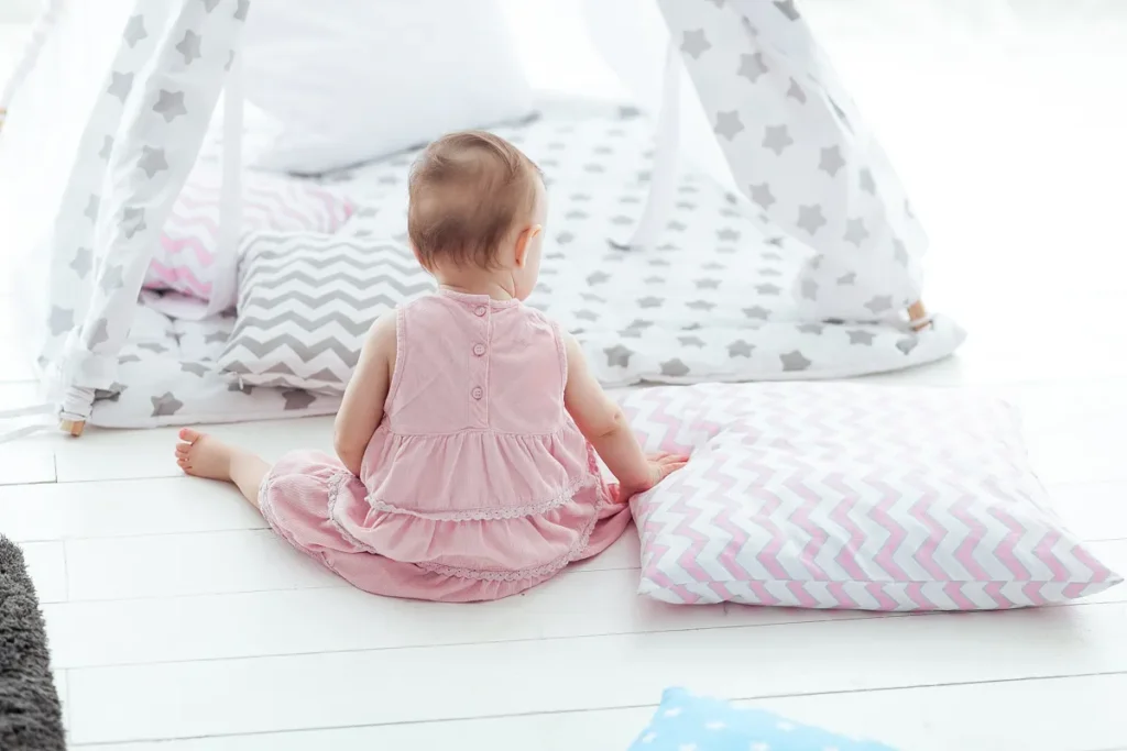 Bebê de vestido rosa sentada de costas sobre um piso de madeira branca, cercada por almofadas firmes estampadas em zigue-zague e uma cabana de brinquedo ao fundo.