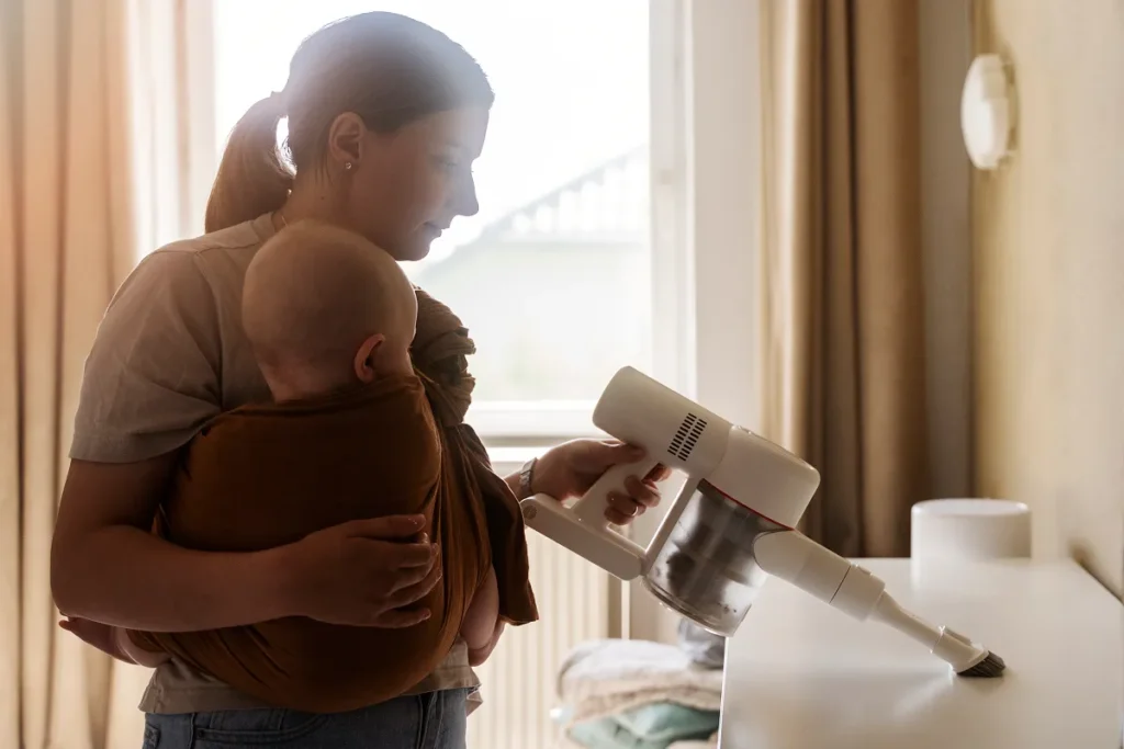 Mãe carregando o bebê em um sling marrom enquanto usa um aspirador de pó portátil sem fio branco para limpar a superfície de um móvel claro.