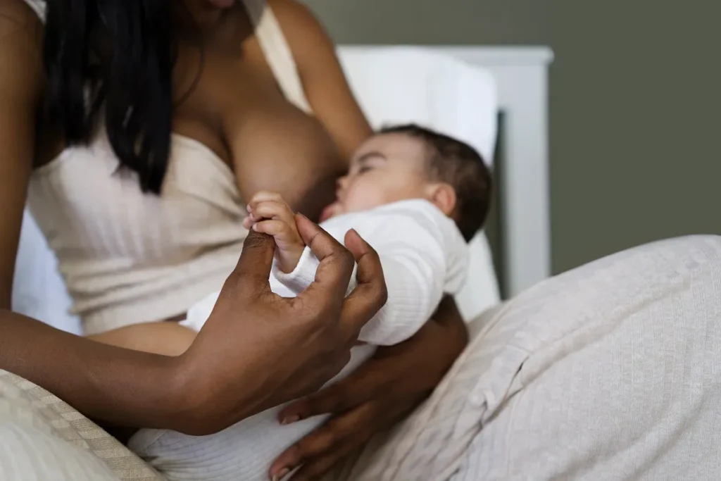 Bebê deitado de bruços em uma cama com lençóis cinzas, com um berço de grades brancas ao fundo.