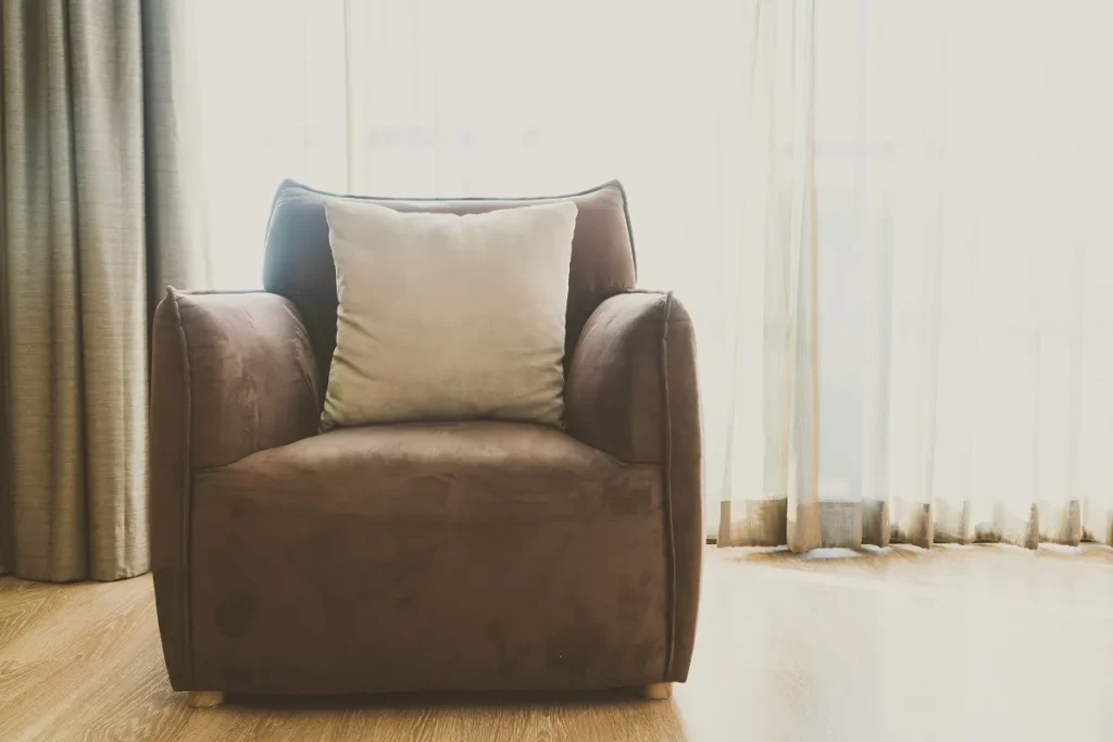 Poltrona de amamentação marrom texturizada com almofada bege clara em quarto com piso de madeira e cortina fina.