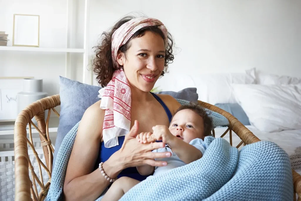 Mãe sorrindo sentada em poltrona de vime com o bebê no colo envolto em manta azul de tricô.