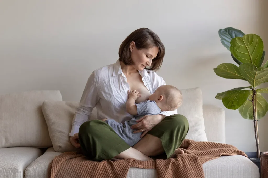Mãe sentada de pernas cruzadas amamentando o bebê no colo em um sofá bege com almofadas ao fundo.