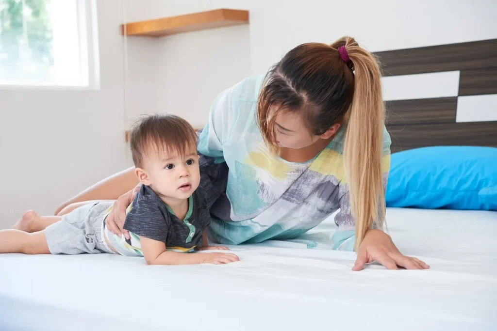 Menina pequena sorridente com marias-chiquinhas no cabelo, vestindo blusa branca e short jeans, sentada em um tapete felpudo cinza dentro de um quarto infantil iluminado com berço ao fundo.