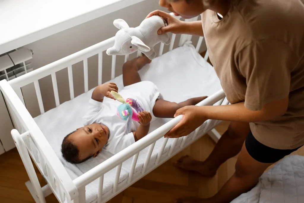 Vista através das grades de madeira de um berço, mostrando um bebê de fralda deitado de costas em um lençol branco, enquanto as mãos de um adulto o amparam delicadamente segurando um brinquedo colorido.