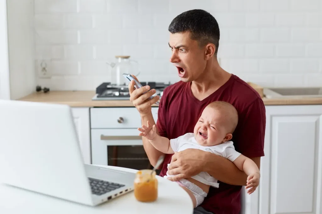 Pai expressando frustração ou surpresa ao olhar para o celular enquanto segura um bebê chorando no colo em uma cozinha, ilustrando a importância do Modo VOX e de alertas eficientes de som.