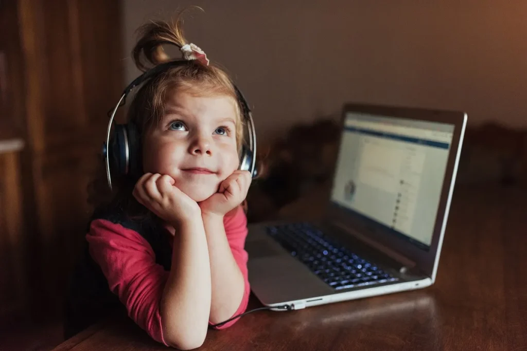 Menina pequena apoiando o queixo nas mãos e usando fones de ouvido grandes enquanto olha para cima e para o lado, sentada ao lado de um notebook aberto em uma mesa de madeira escura.