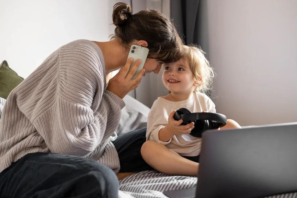 Mãe de perfil falando ao celular e encostando a testa na de sua filha pequena, que está sentada na cama sorrindo com fones de ouvido nas mãos em frente a um notebook aberto.