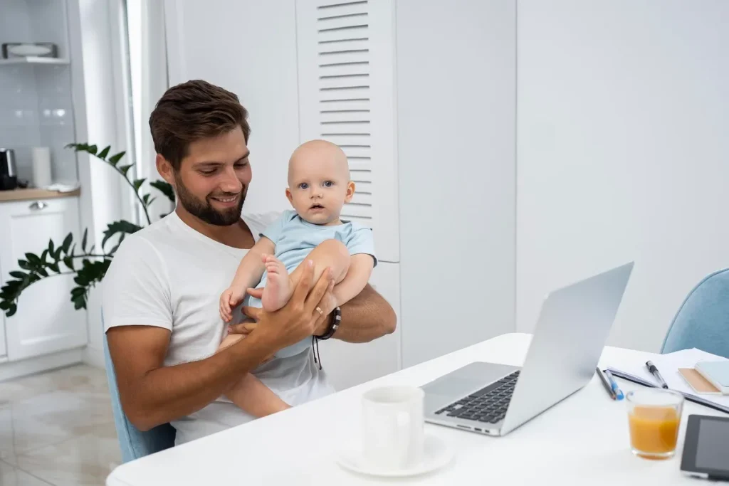 Pai sentado em uma cadeira segurando um bebê no colo à mesa, enquanto olha sorrindo para o filho ao lado de um laptop aberto, ilustrando o planejamento de um ambiente seguro e sem fios soltos.