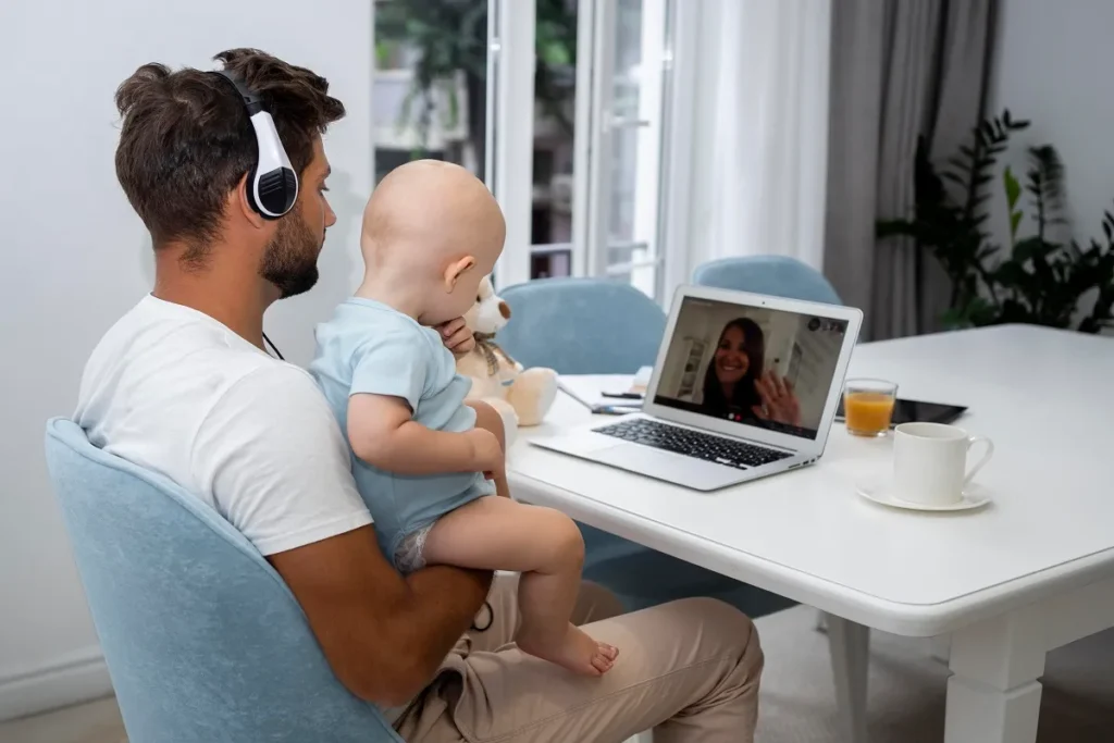 Pai usando fones de ouvido grandes e segurando um bebê no colo enquanto faz uma videochamada no laptop com uma mulher na tela, ilustrando o uso de dispositivos sem fio em uma mesa de jantar branca.