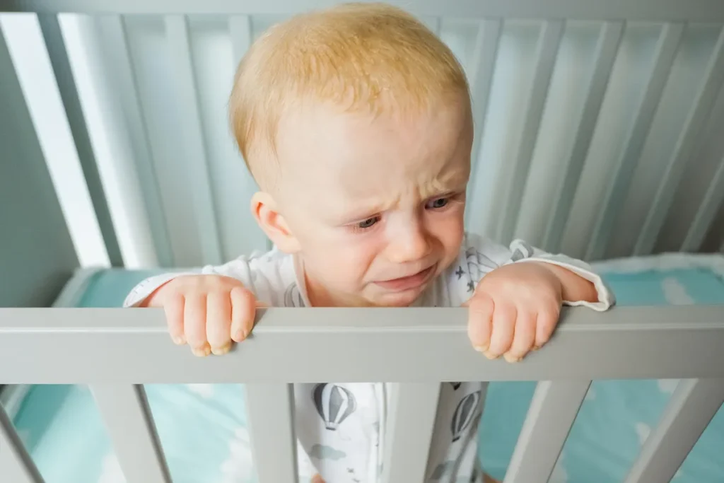 Close de um bebê loiro chorando com expressão de angústia, em pé dentro de um berço cinza, segurando firmemente a grade superior com as duas mãos.