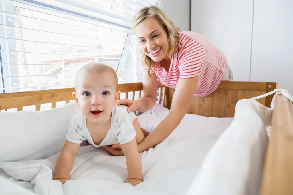 Mãe loira sorrindo e amparando o bebê de macacão branco que está de bruços, engatinhando sobre um colchão macio dentro de um berço de madeira clara.
