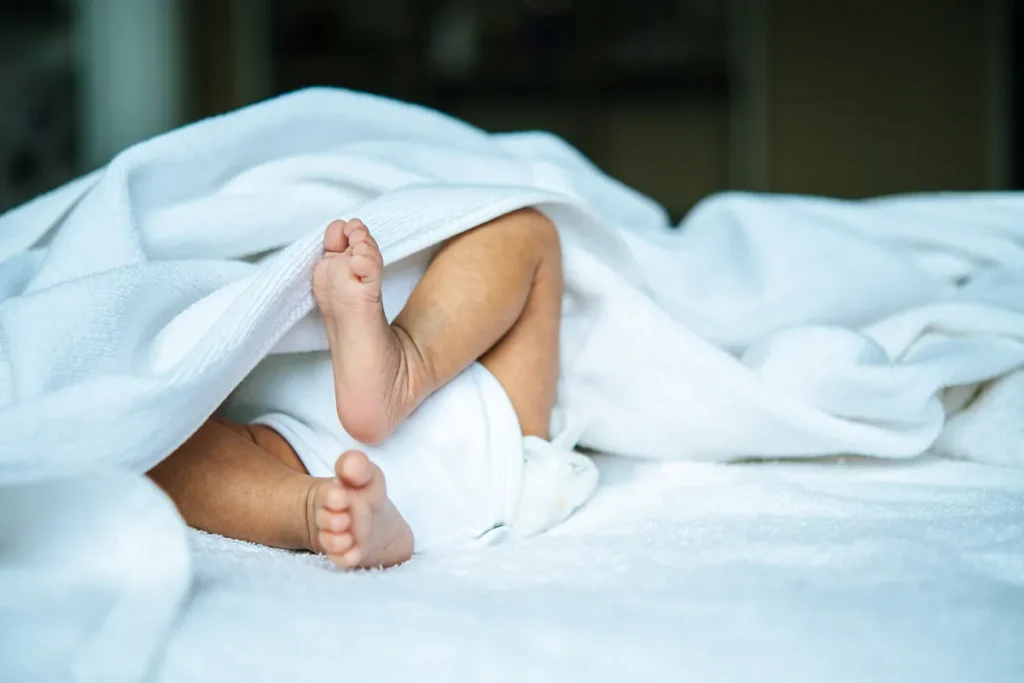 Close dos pezinhos e pernas de um bebê recém-nascido deitado de costas, parcialmente coberto por uma manta branca espessa e fofa que envolve seu corpo.