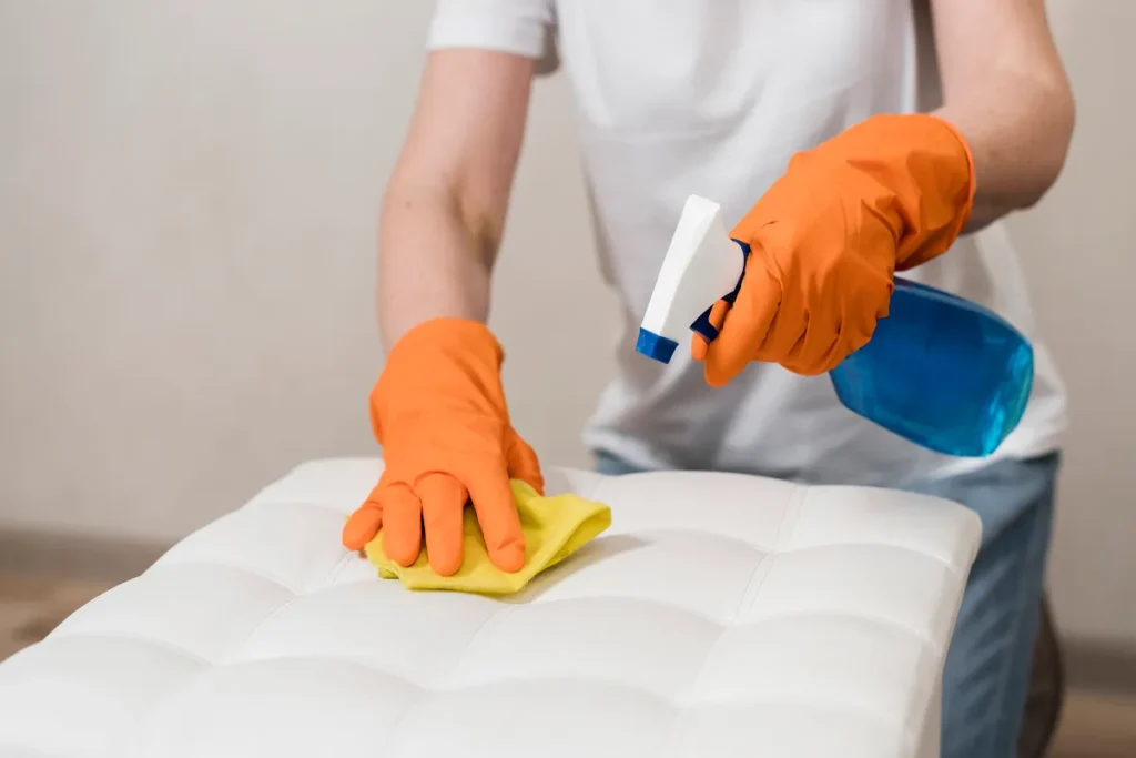 Close das mãos de uma pessoa usando luvas laranjas, segurando um borrifador azul com spray e esfregando um pano amarelo sobre um estofado branco textuado.