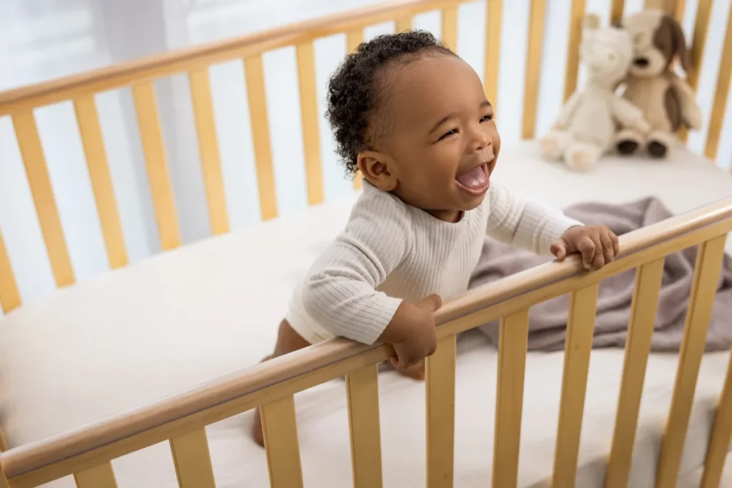 Bebê sorrindo em pé dentro de berço portátil com estrutura elevada e ambiente infantil iluminado.