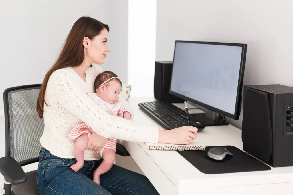 Mãe sentada em uma cadeira de escritório segurando um bebê no colo enquanto trabalha em uma mesa com computador de mesa e anota em um caderno, representando a proteção da rede doméstica para a segurança da família.