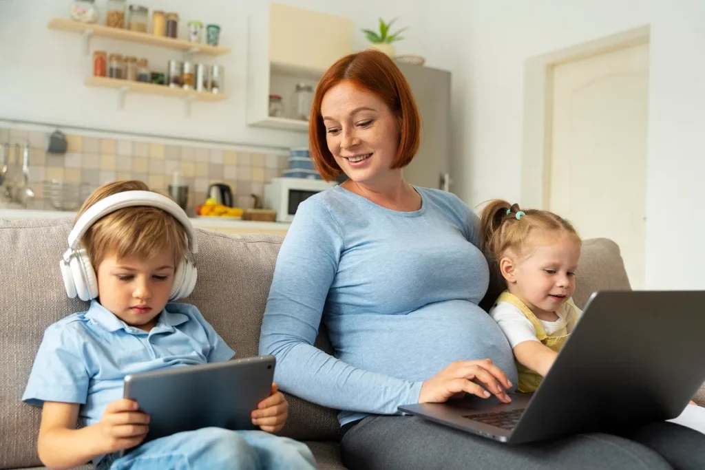 Mãe grávida sentada no sofá usando um notebook com a filha pequena ao lado, enquanto seu filho usa um tablet com fones de ouvido, ilustrando o alto consumo e interferência na rede residencial Wi-Fi de 2.4GHz e 5GHz.