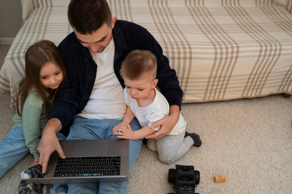 Pai sentado no chão com um menino e uma menina usando um notebook, representando o cuidado com telas e a manutenção de aparelhos portáteis e eletrônicos de monitoramento em casa.