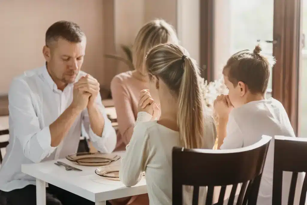 Família com pai, mãe e dois filhos orando juntos à mesa antes do almoço em ambiente iluminado