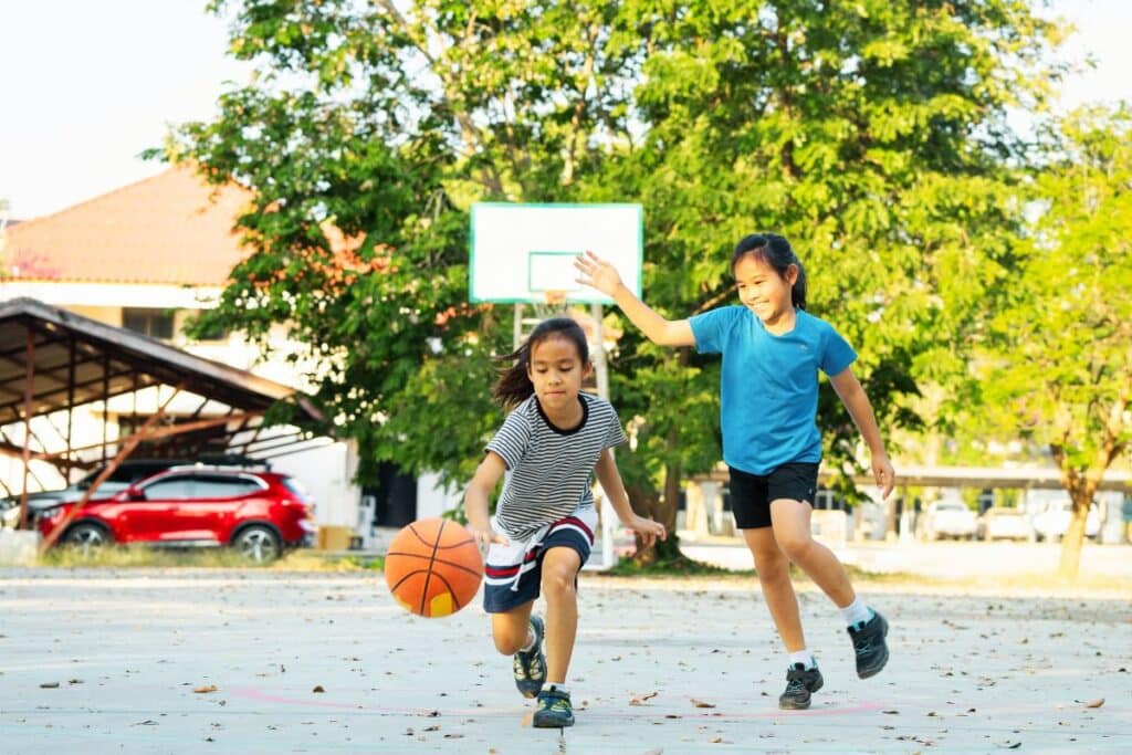 Duas crianças brincando com bola de basquete em quadra ao ar livre, representando brinquedo para criança de 7 anos que estimula movimento e socialização.