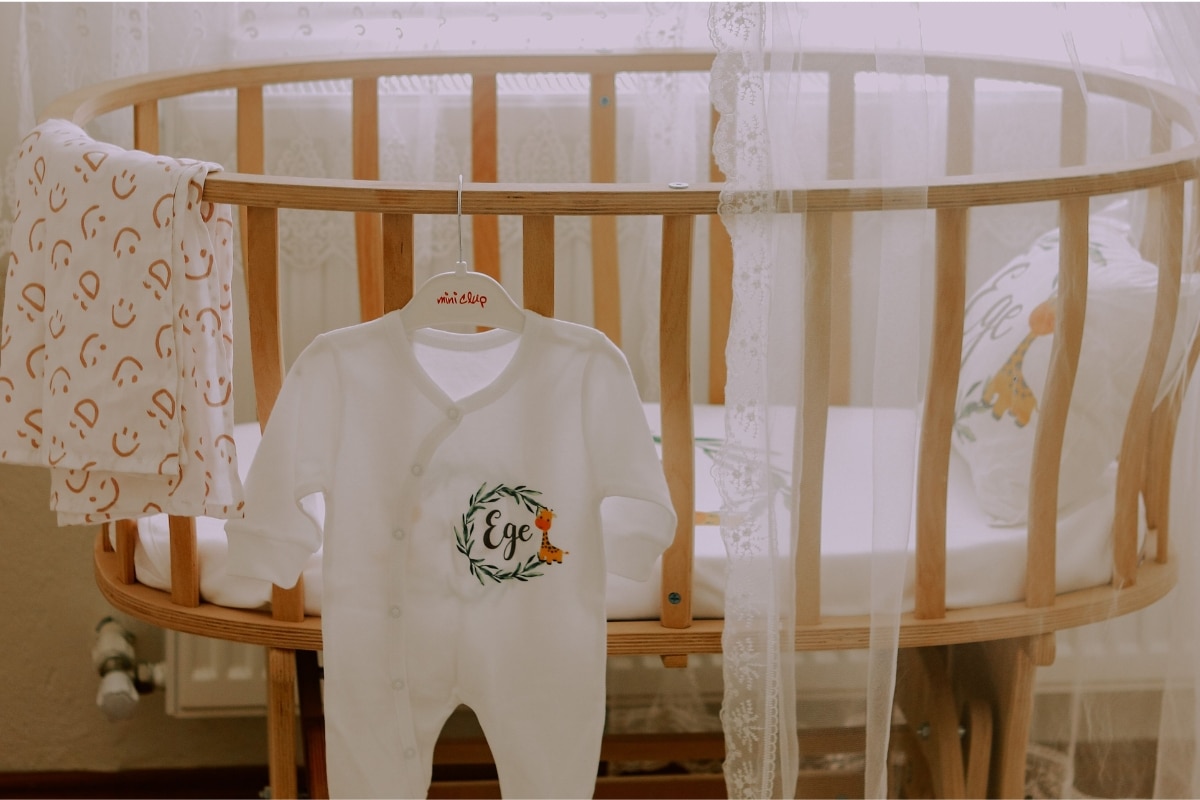 Mini berço oval de madeira com roupas de bebê e dossel em um quarto infantil.