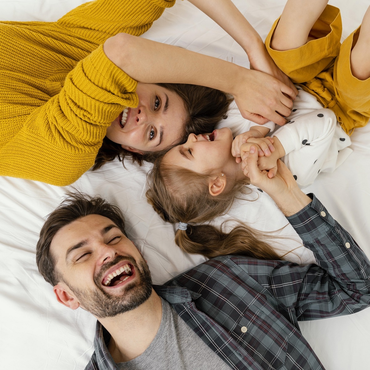 um homem, uma mulher e uma criança loira deitados em um lençol branco rindo e brincando