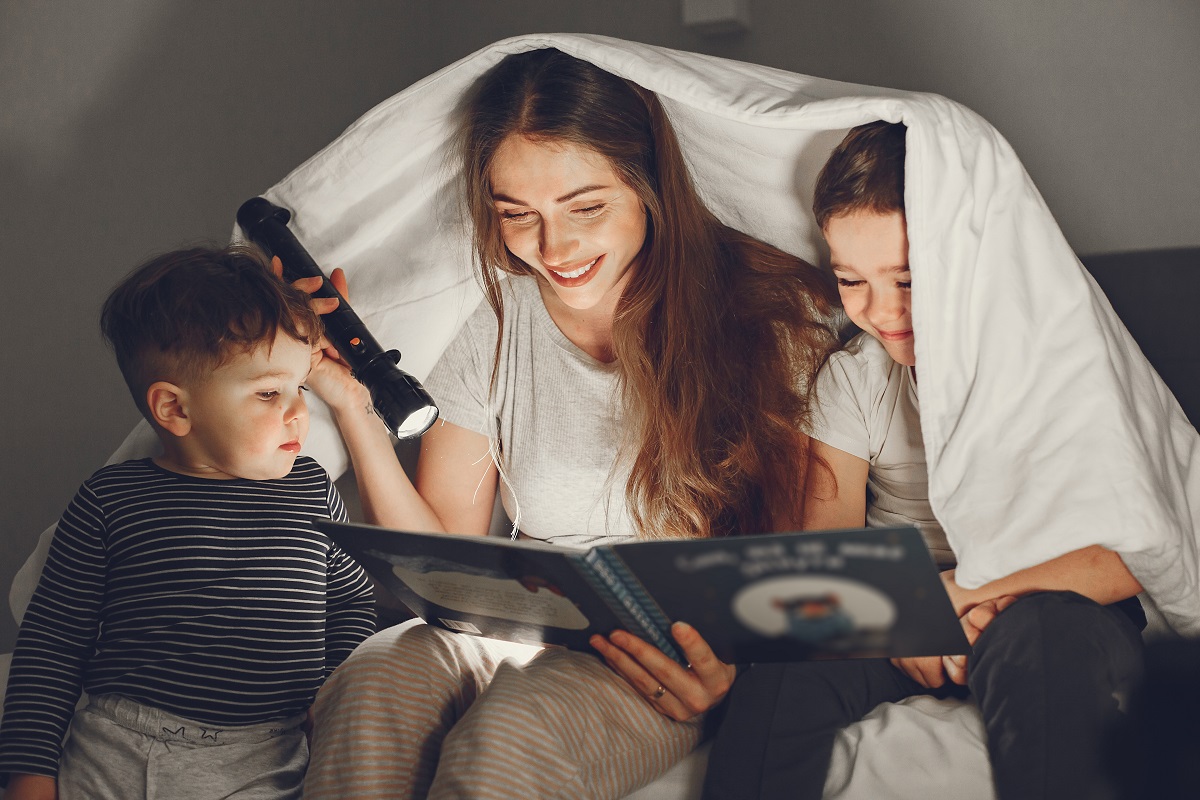 uma mulher entre duas crianças, um menino e uma menina, os três estão lendo um livro, a mulher está iluminando o livro com uma lanterna acesa