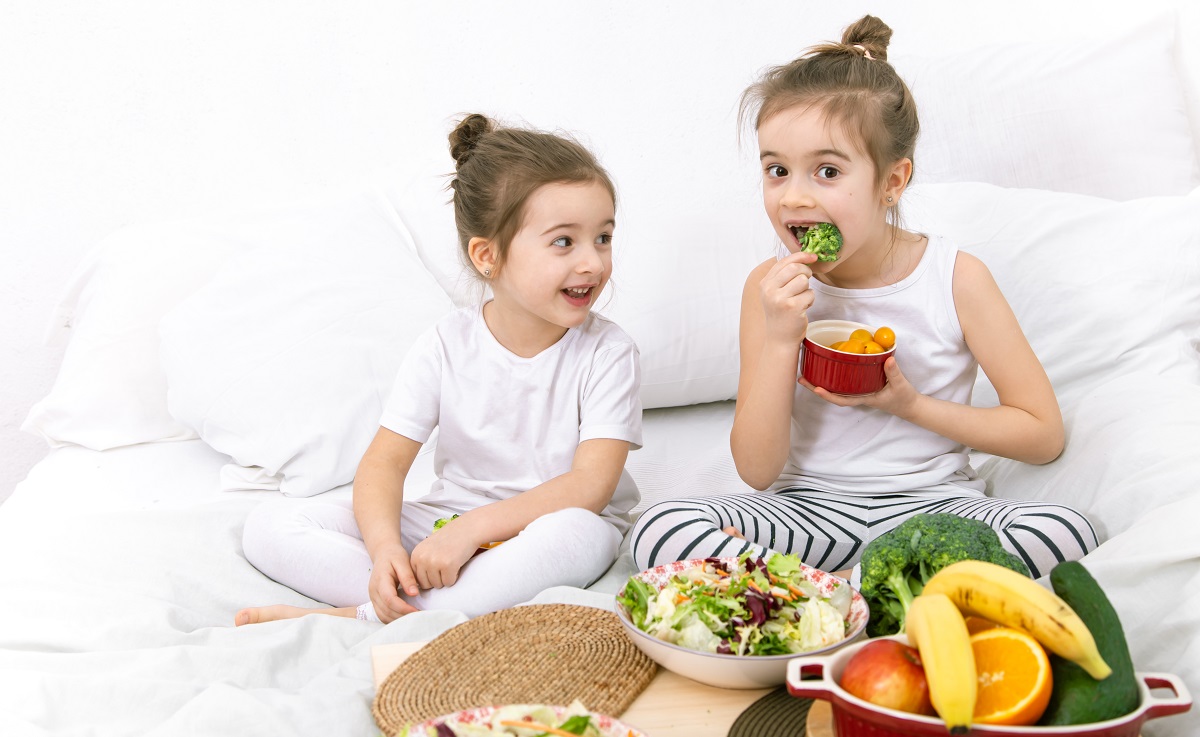 duas meninas sentadas em uma cama comendo verduras