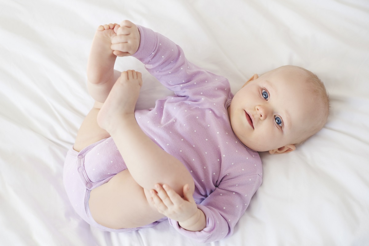 bebê deitado com as pernas para cima vestindo uma camisa roxa de bolinhas brancas