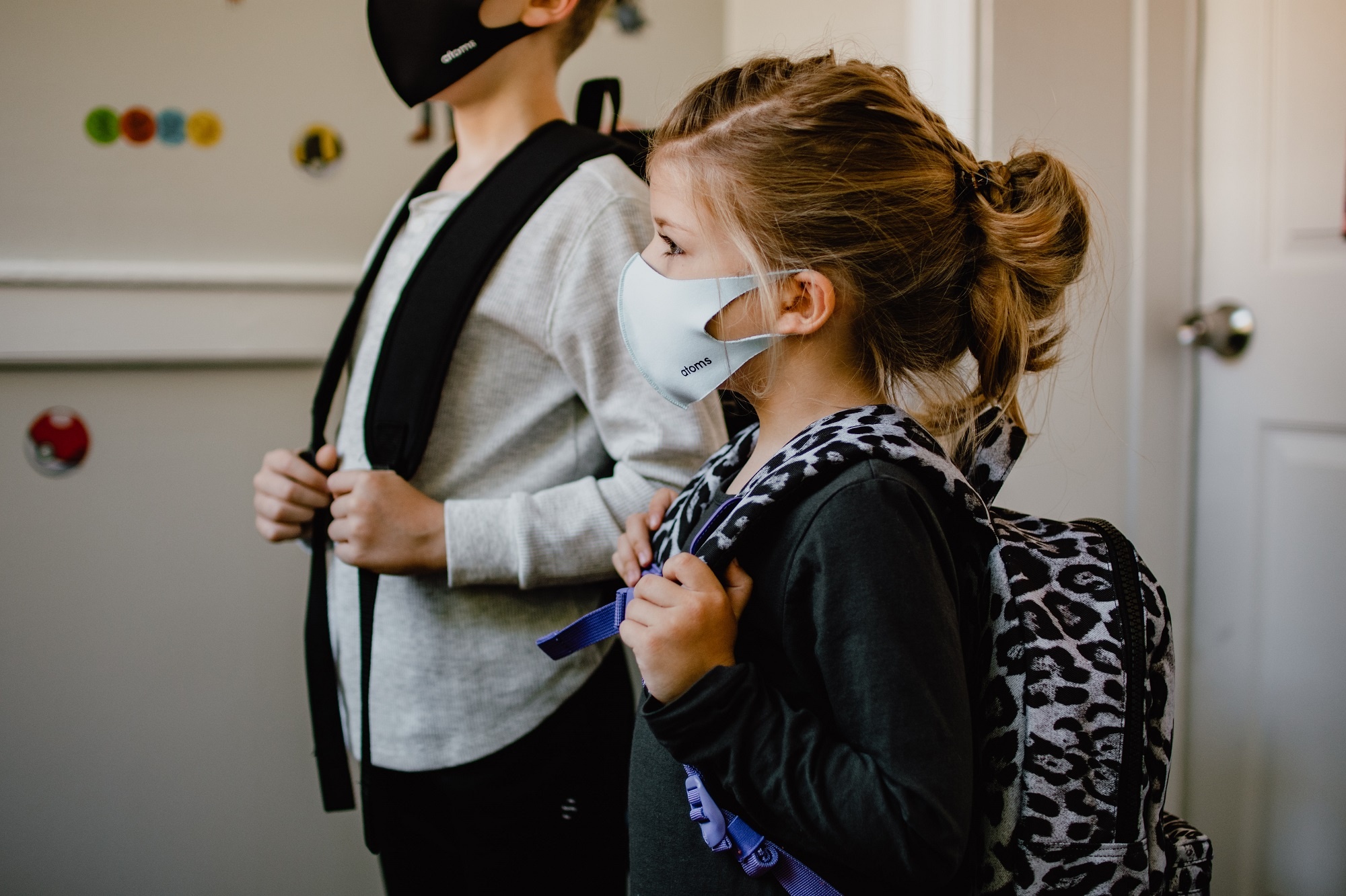uma menina e um menino de pé lado a lado usando máscaras no rosto com mochilas nas costas