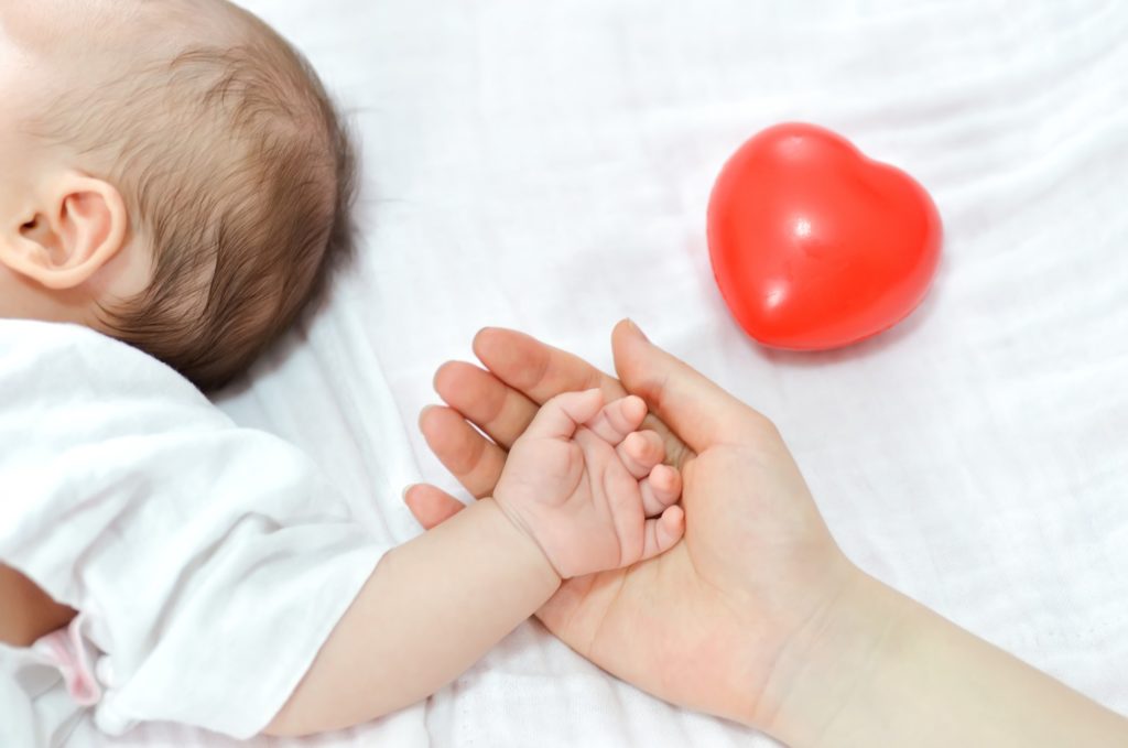 mão feminina adulta segurando a de um bebê com um coração vermelho mais ao fundo