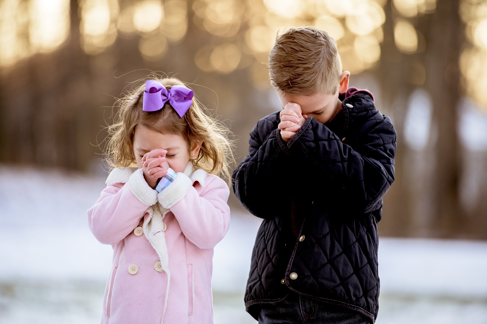 uma menina loira pequena e um menino loiro pequeno rezando em um jardim coberto de neve com um fundo desfocado