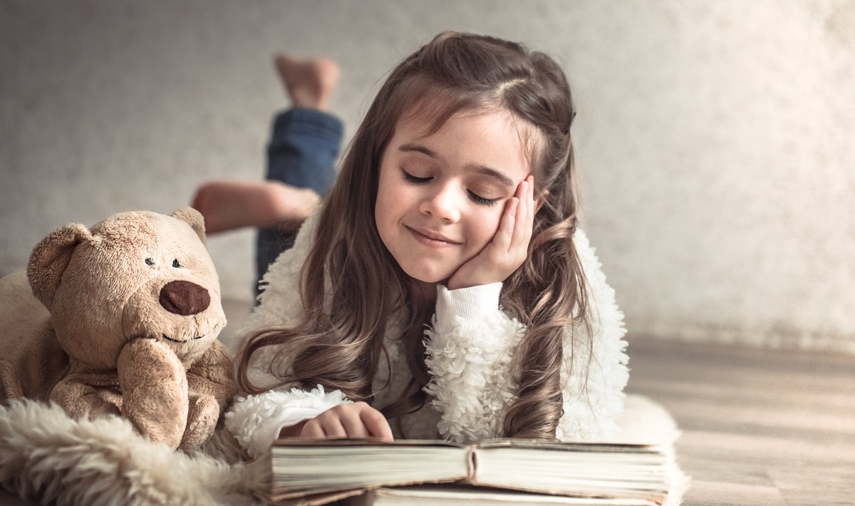 menina sonhadora deitada de braços no chão com a mão sobre um livro e um urso de pelúcia do lado