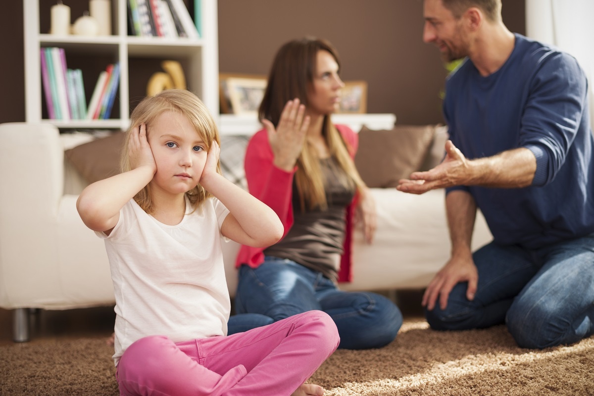 um homem e uma mulher no fundo desfocados parecem estar discutindo uma menina no primeiro plano loira está tampando os ouvidos