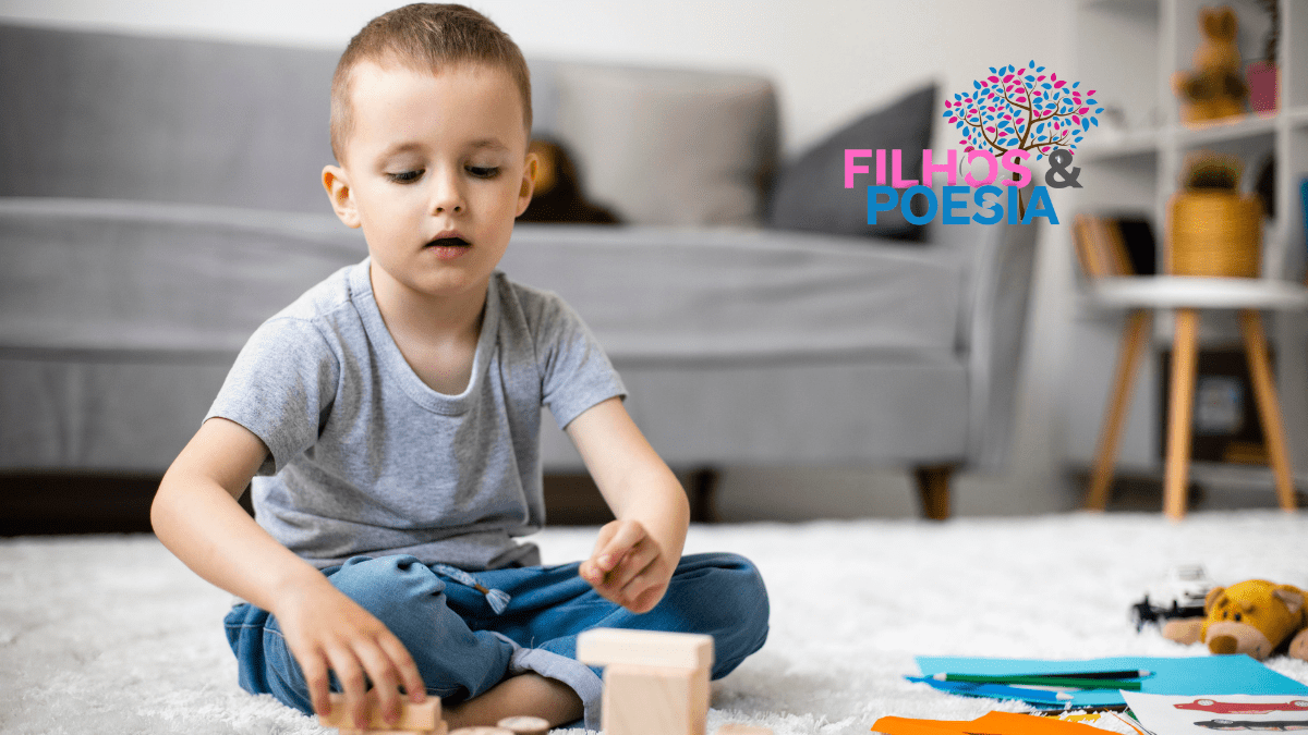 menino sentado na sala de estar brincando com blocos de madeira vestindo calça jeans e camisa cinza simples