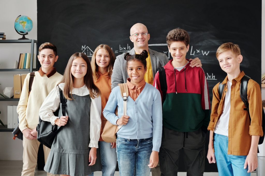 professor e seus alunos de pé em frente ao quadro negro na sala de aula todos sorrindo juntos