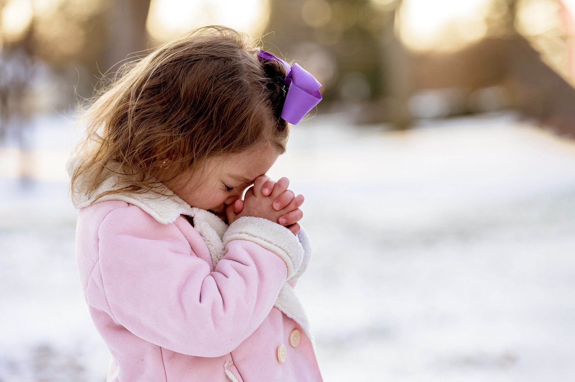 Menina loira rezando em um jardim coberto de neve sob a luz do sol a uma distância borrada
