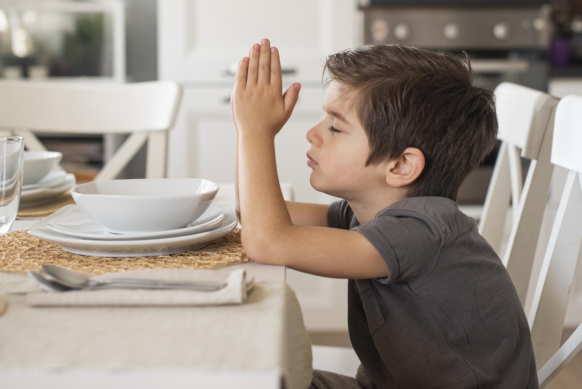 menino sentado a mesa de olhos fechados rezando