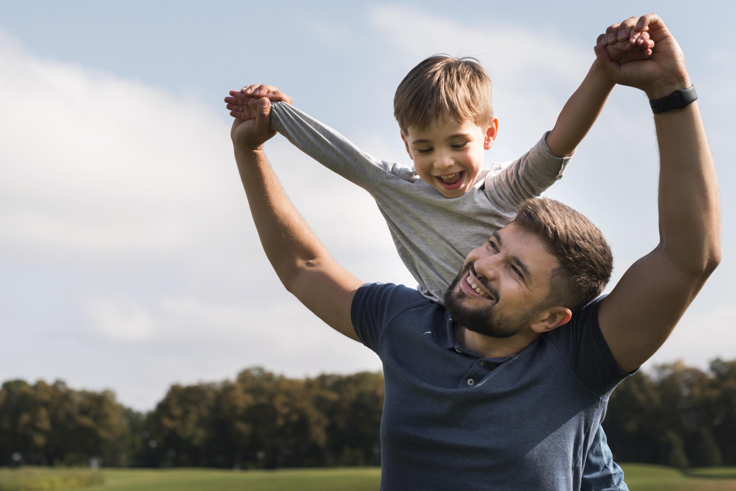 homem jovem adulto com um menino nas costas o segurando pela mão ambos estão sorrindo em um parque